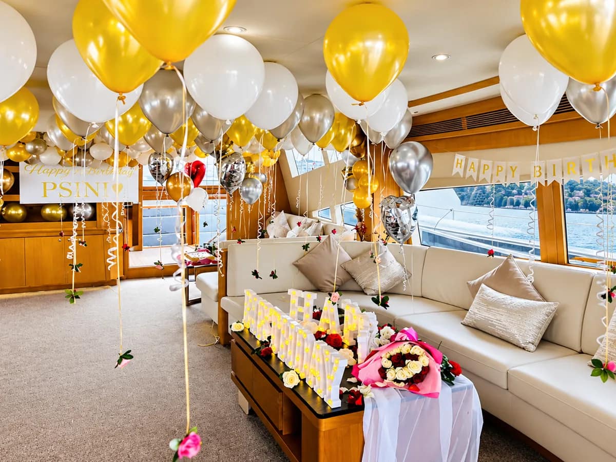 Yachtsalon dekoriert mit goldenen, weißen und silbernen Luftballons, einem HAPPY-BIRTHDAY-Banner, Leuchtbuchstaben und einem Rosenstrauß